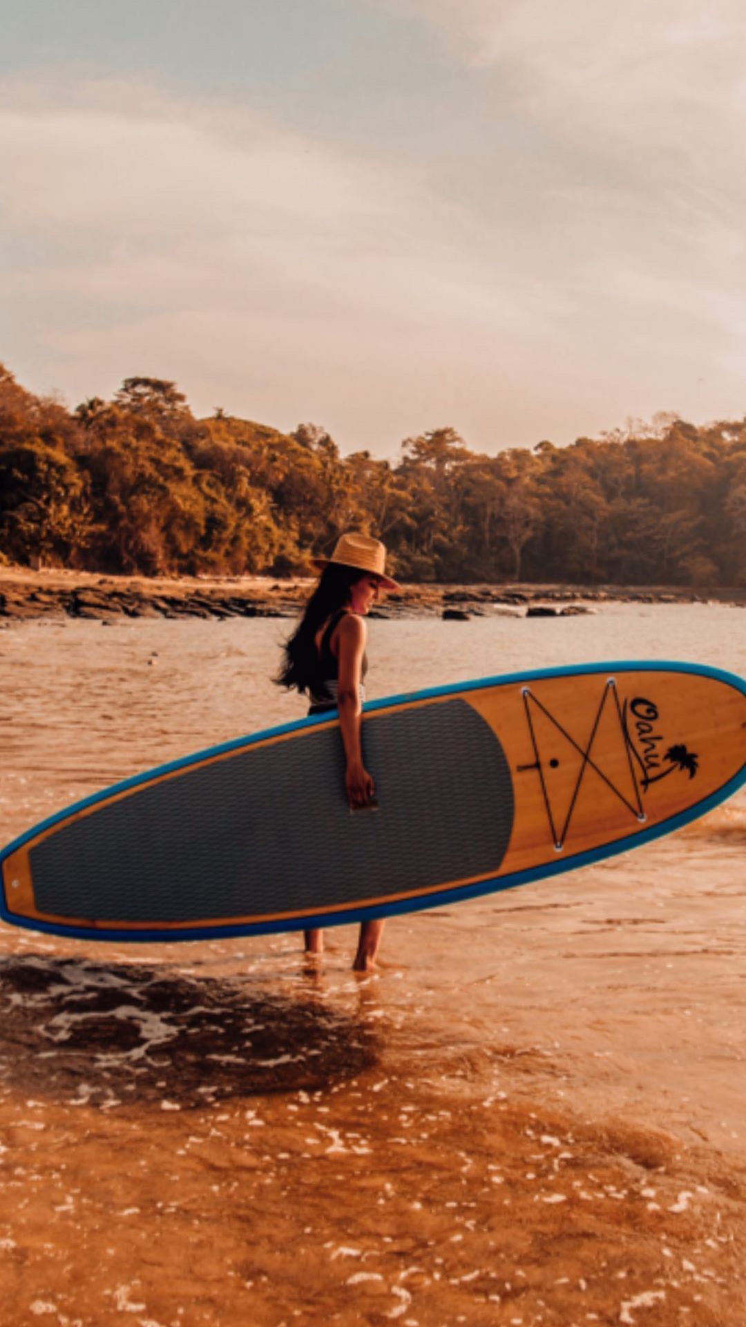 A person holding a surfboard with a logo, standing in shallow water with a beach and trees in the background.