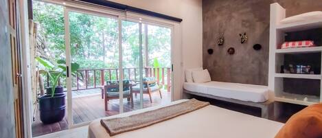 A bedroom with a large bed, a sofa, a wooden deck with chairs, and a view of greenery through large windows.