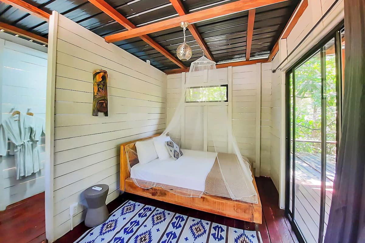 Une chambre avec un lit en bois, une moustiquaire, une petite fenêtre et un tapis à motifs.
