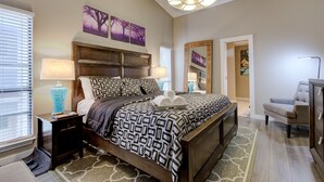Une chambre avec un grand lit, une table de chevet, une commode et une chaise. On y trouve également des œuvres d’art murales et un luminaire au plafond.
