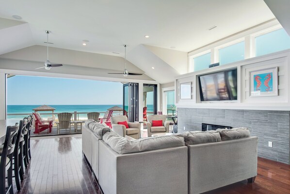 A modern living room with a large sofa, a TV mounted on the wall, and a view of the ocean. - Go Your Own Wave: Oceanfront, Pool, Spa, Elevator (Nags Head)