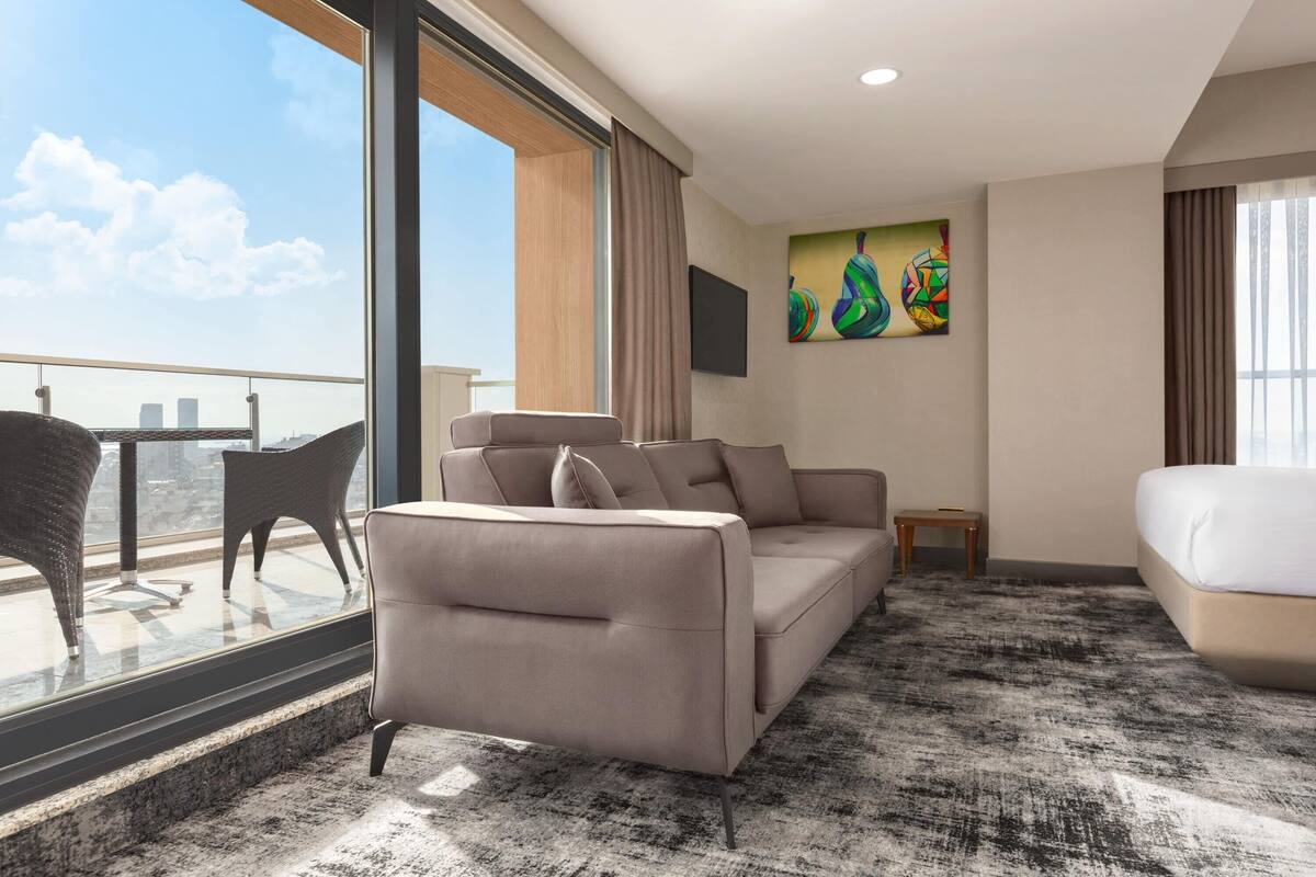 A modern hotel room with a large window, a sofa, a bed, and a small table.