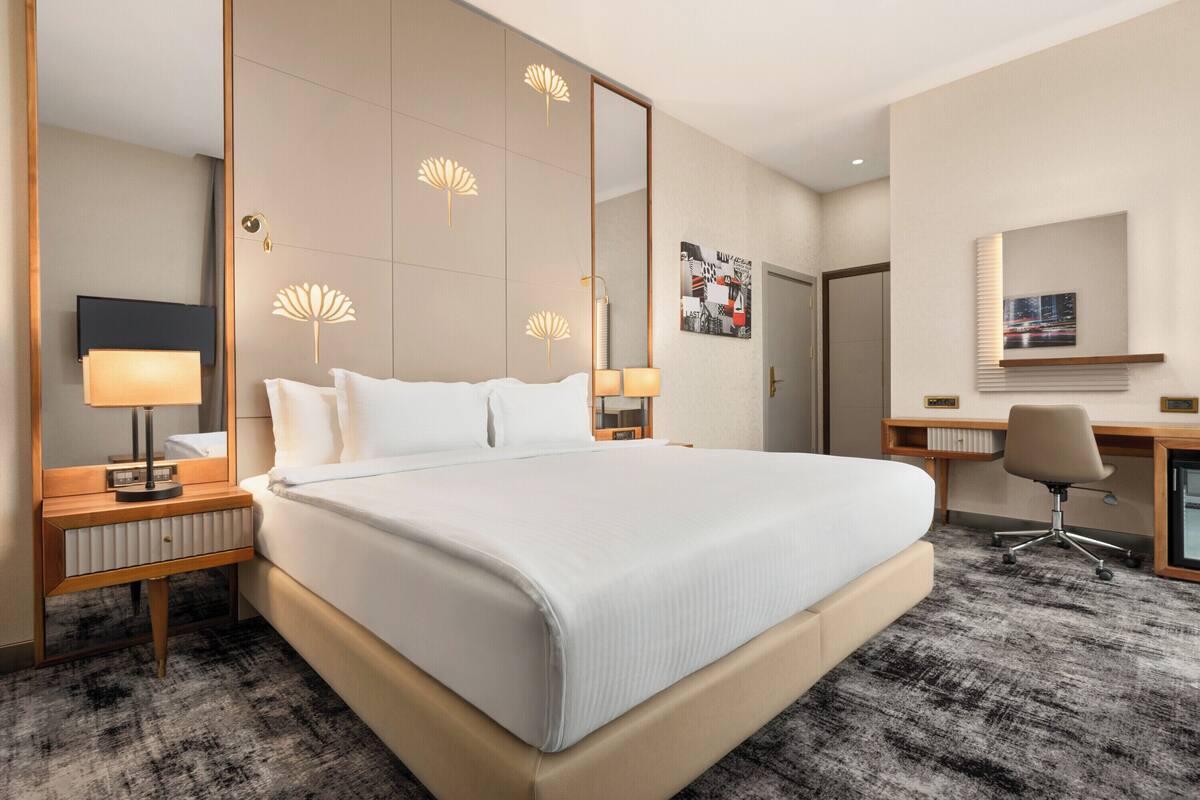 A hotel room with a large bed, bedside lamps, a desk with a chair, a mirror, and a wall-mounted artwork.