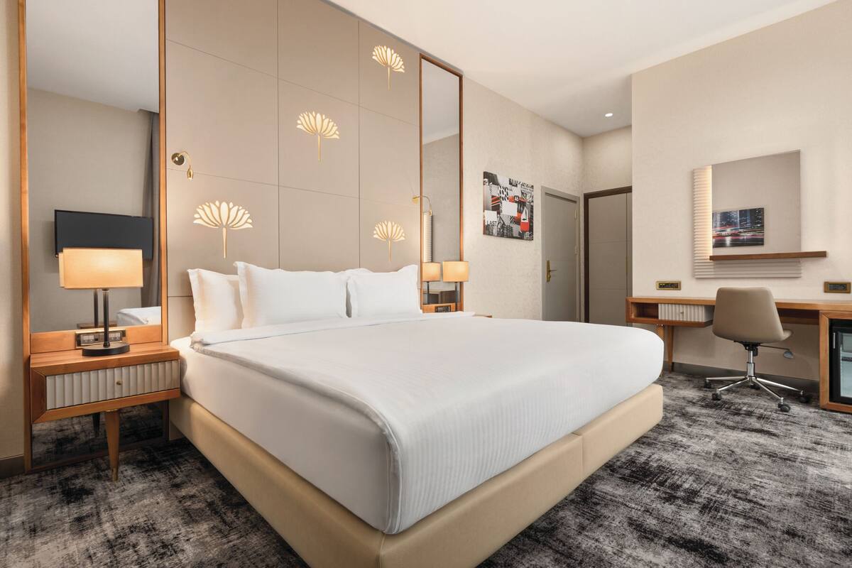 A hotel room with a large bed, bedside lamps, a desk with a chair, a mirror, and decorative wall panels.