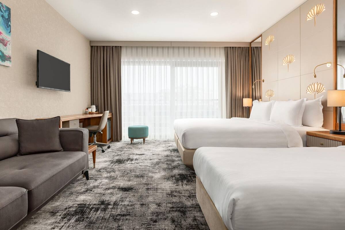 A hotel room with two beds, a sofa, a desk, a TV, and a large window with curtains.