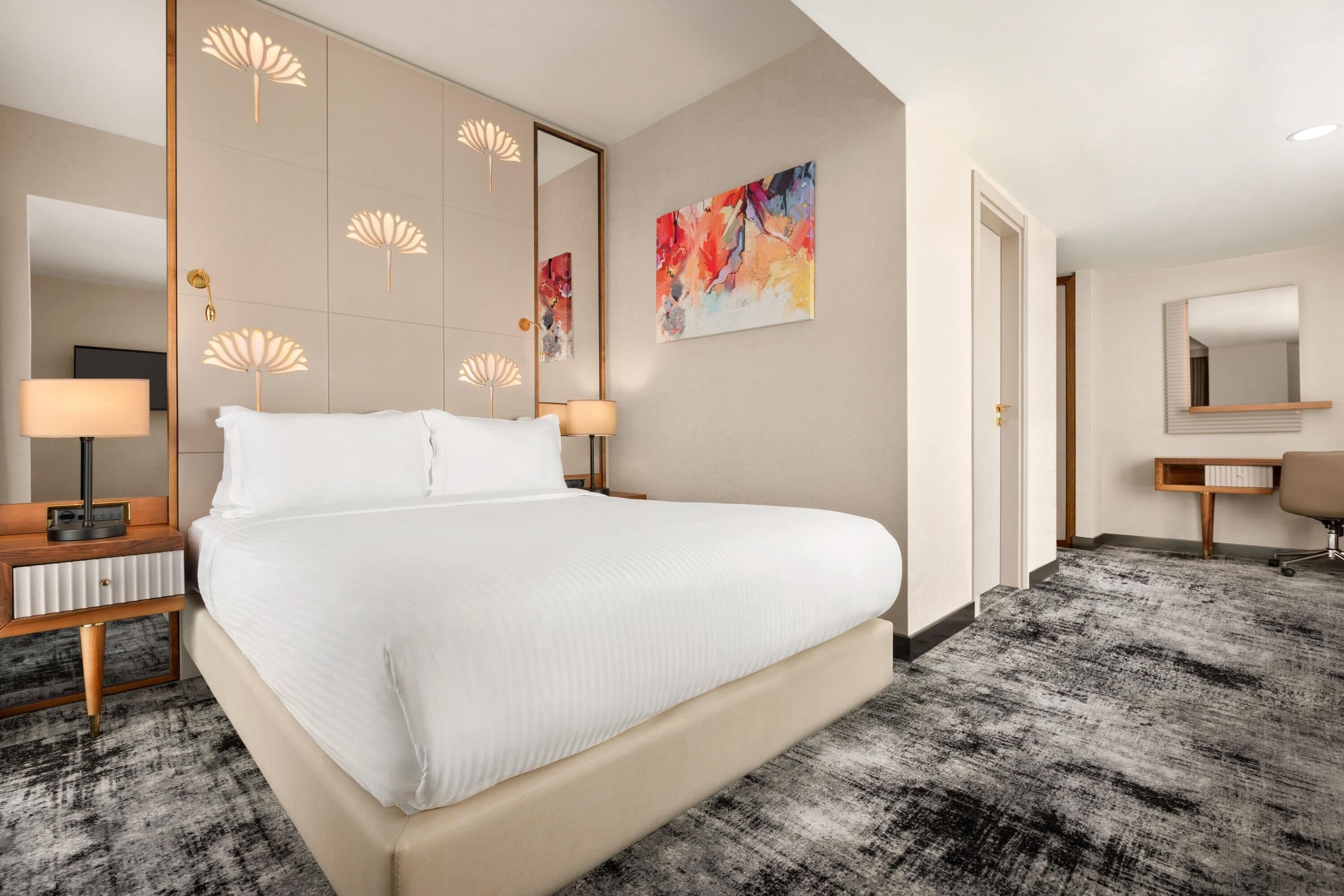 A hotel room with a large bed, bedside lamps, a mirror, a painting, and a carpeted floor.