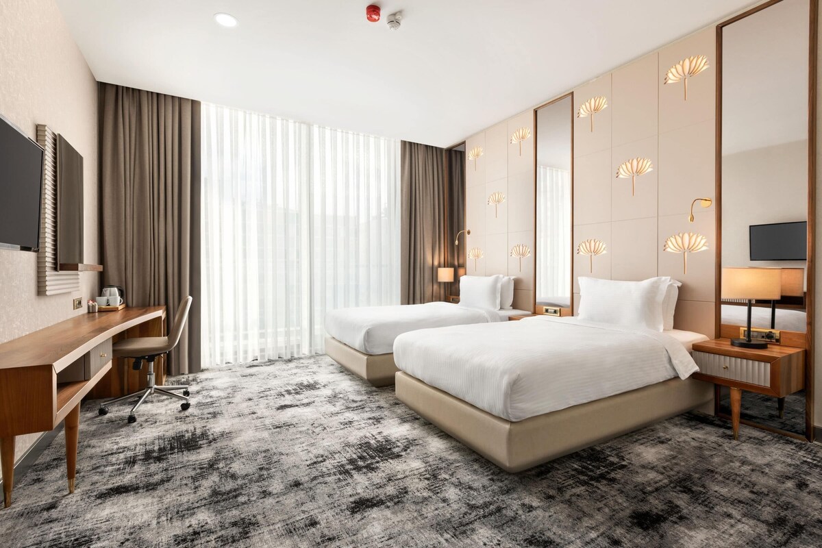 Quarto de hotel com duas camas, uma mesa, uma cadeira, uma televisão e uma janela grande com cortinas.