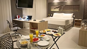 Habitación de hotel con cama, escritorio con una computadora portátil, televisor y mesa de desayuno con diversos alimentos y bebidas.