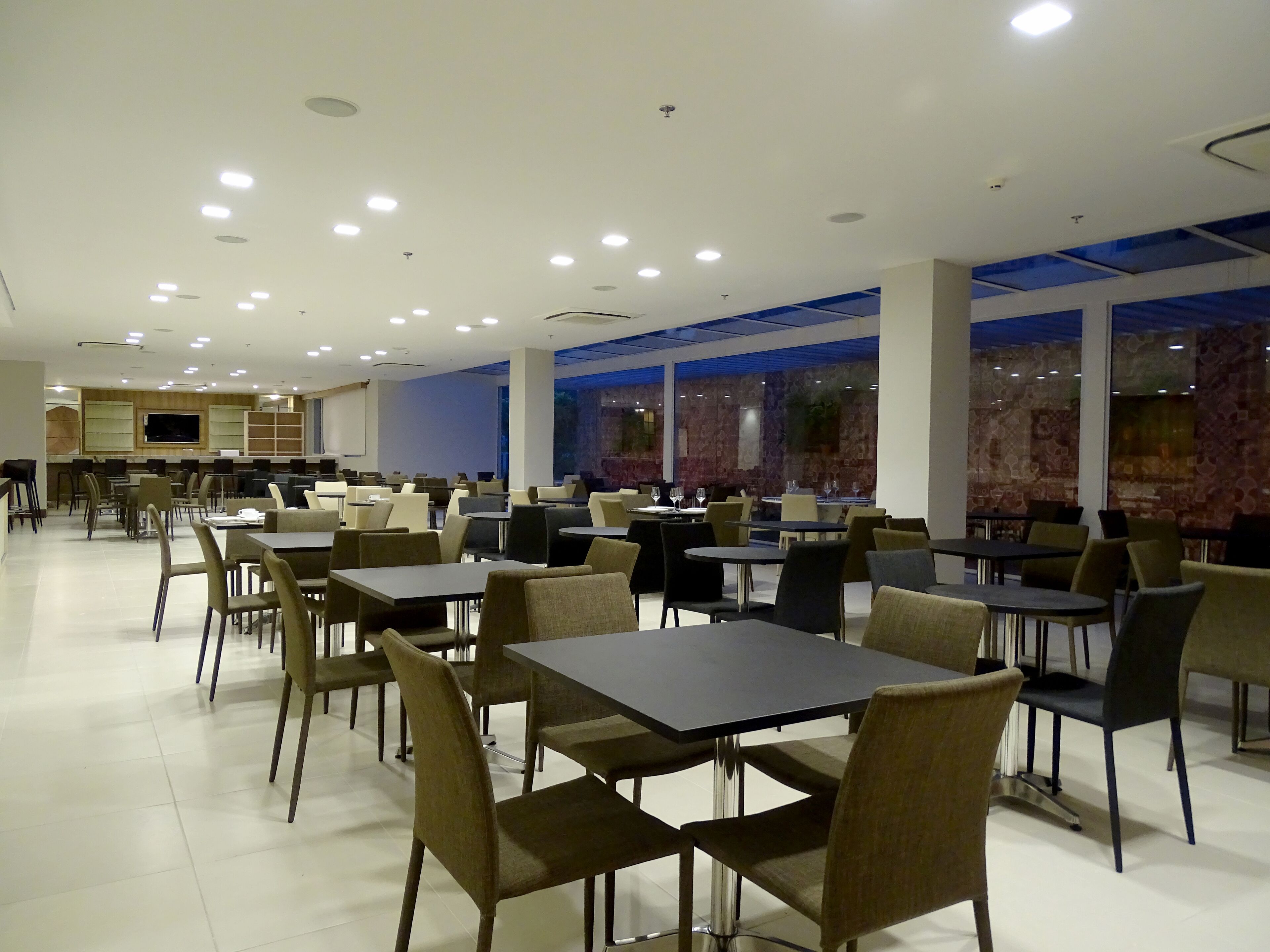 Um restaurante amplo com várias mesas e cadeiras, área de cozinha ao fundo, e grandes janelas que permitem a entrada de luz natural.