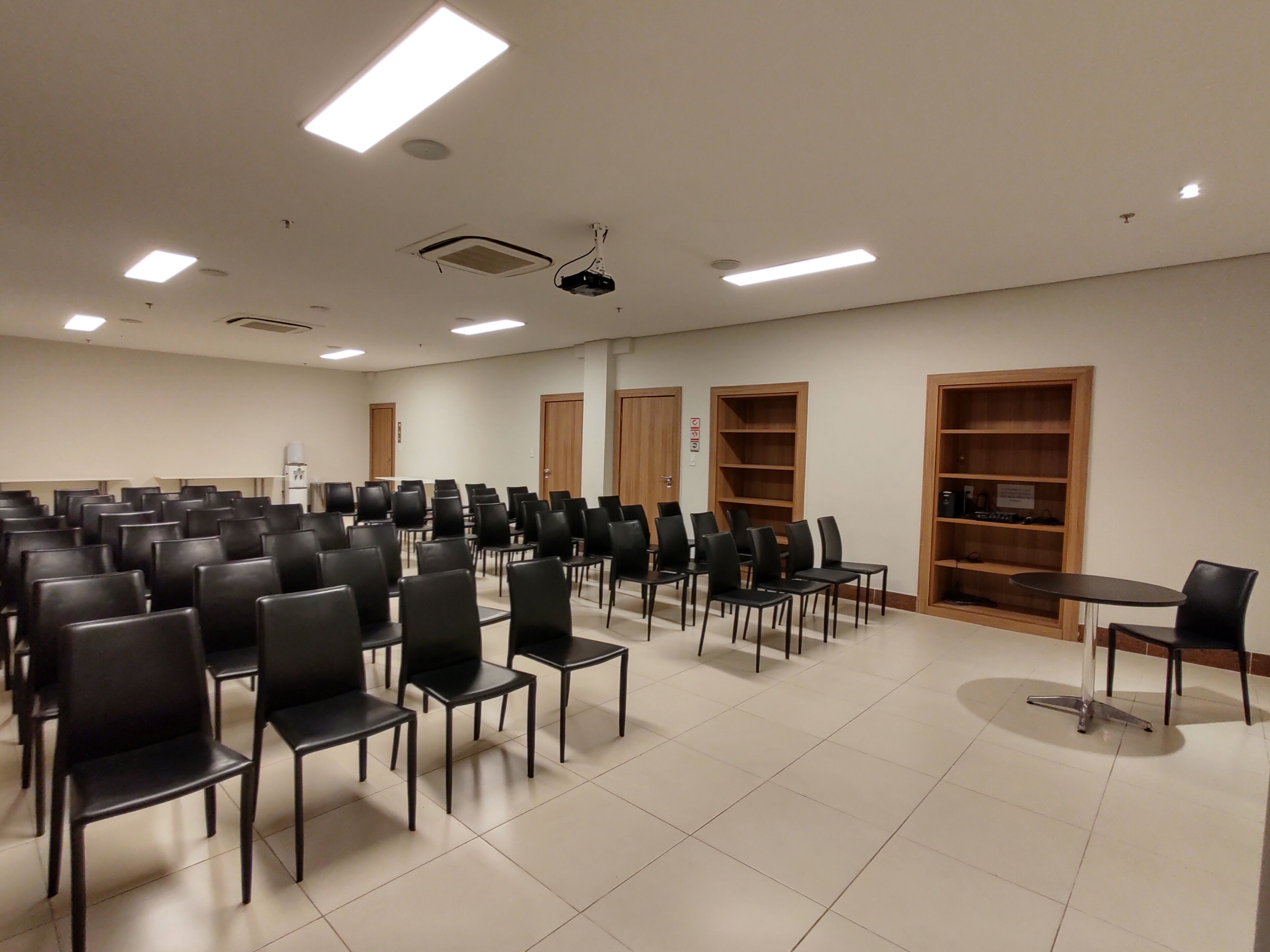 Sala de conferência com fileiras de cadeiras pretas, um projetor e estantes de madeira.