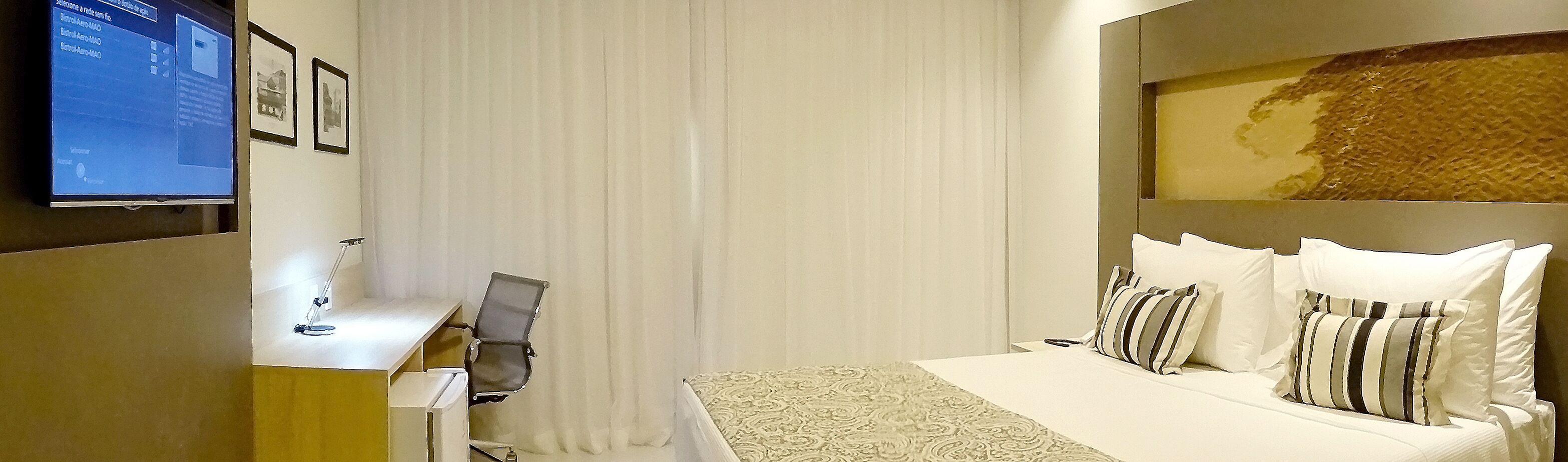 Een hotelkamer met een bed, een bureau met een computer, een televisie en een raam met gordijnen.