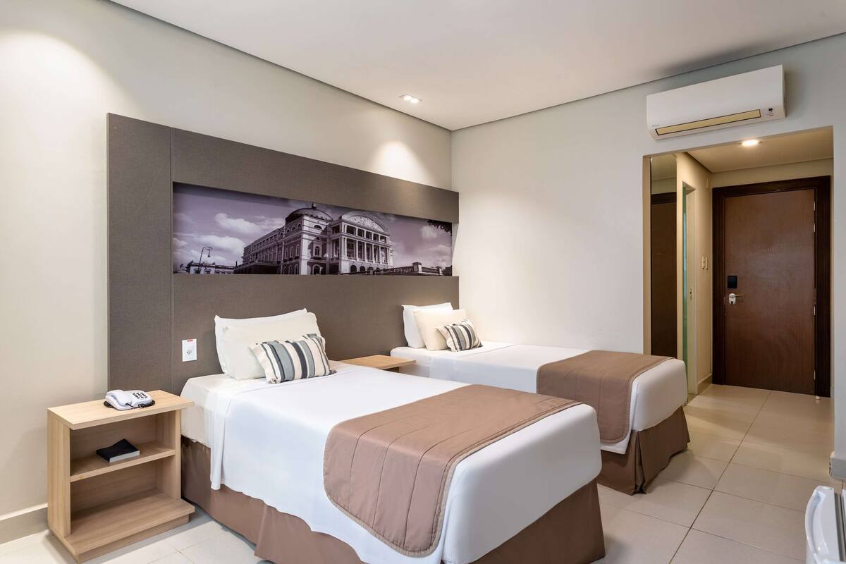 A hotel room with two beds, a bedside table with a phone, a shelf, and a wall-mounted air conditioner.