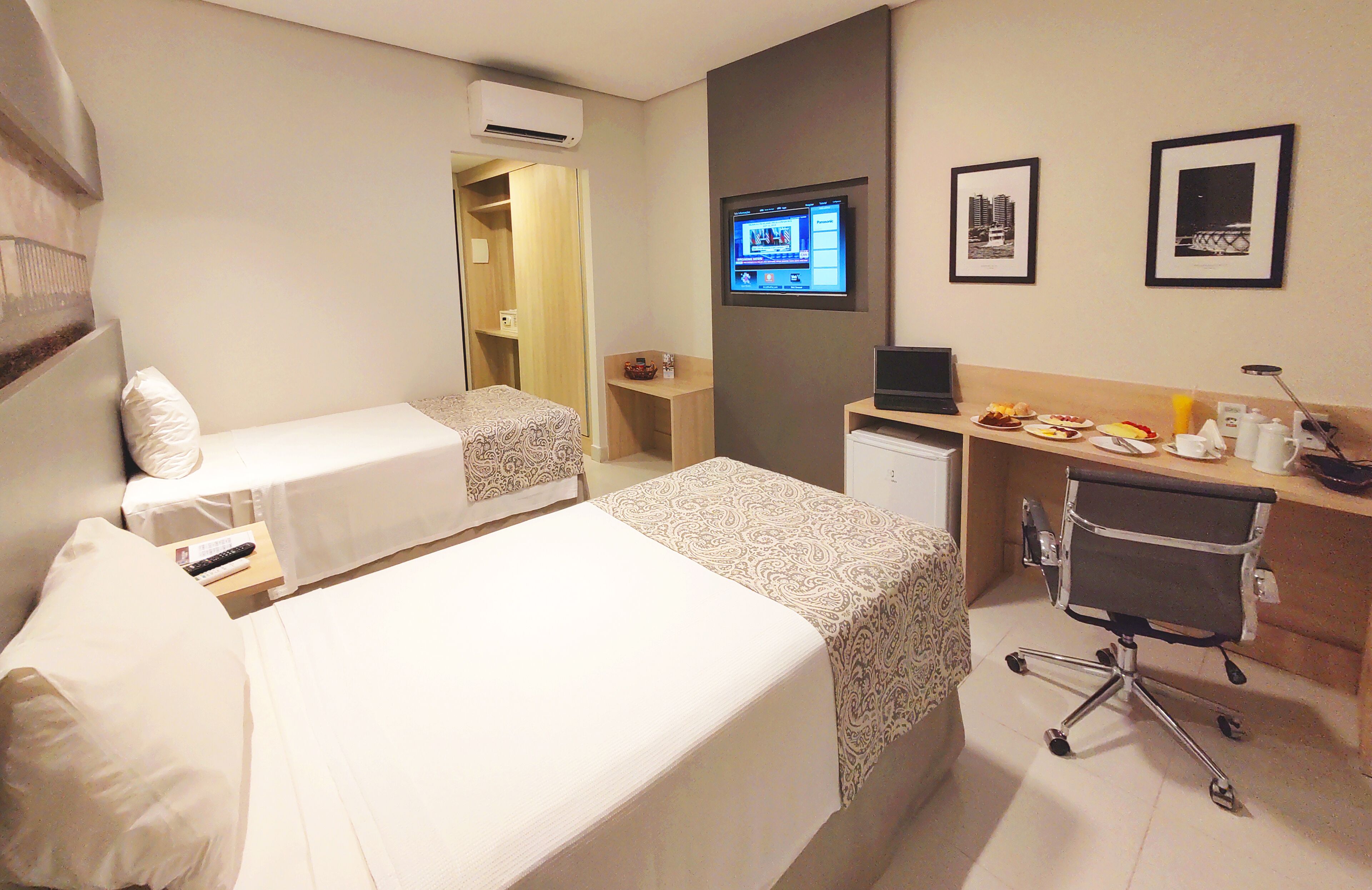 Et hotelværelse med to senge, et skrivebord med computer, en stol og et fjernsyn på væggen.