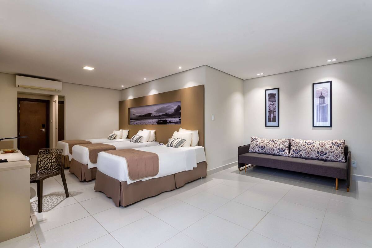 A modern hotel room with two beds, a sofa, a desk, and a large window.