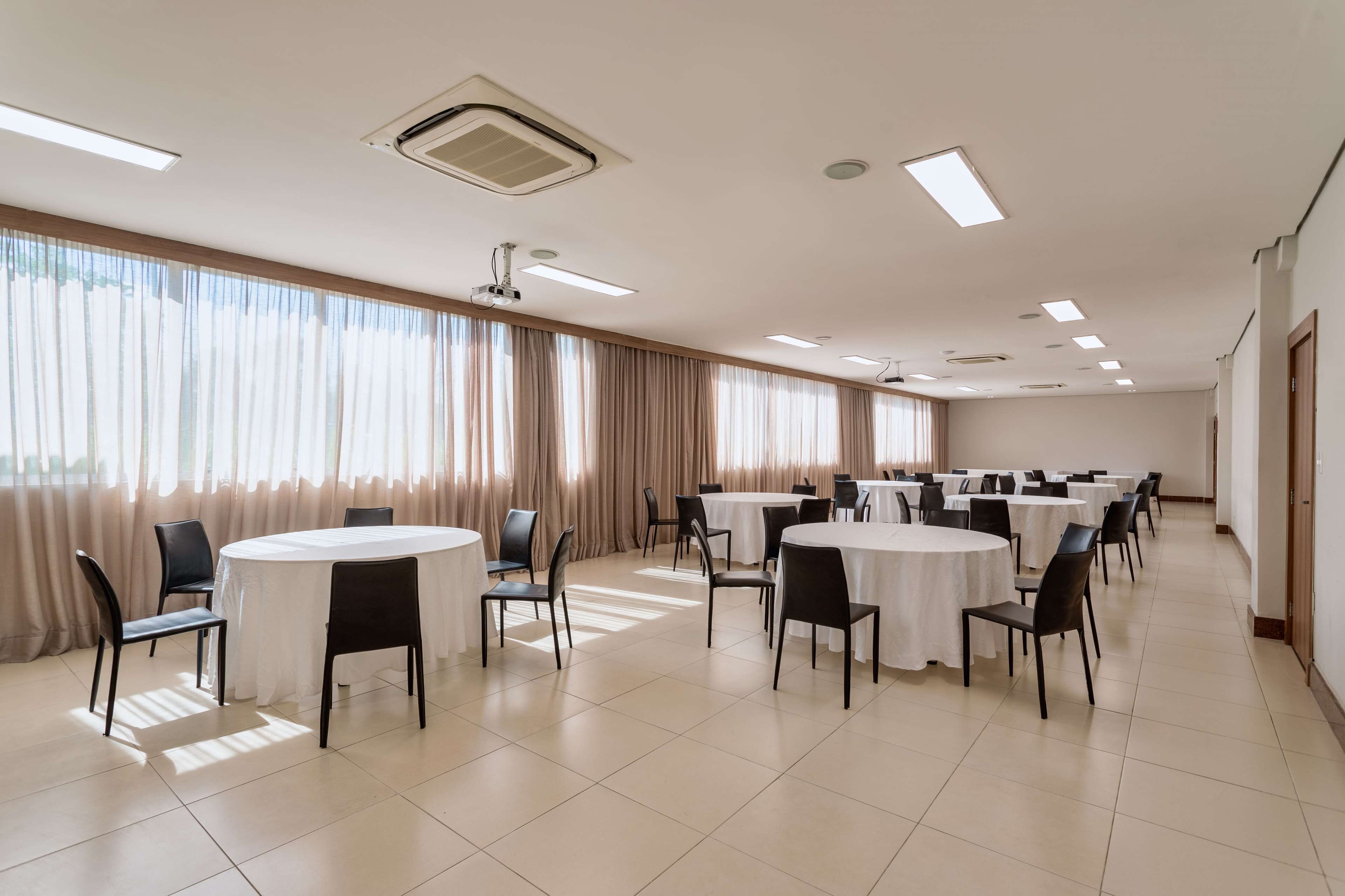A conference room with multiple round tables and chairs, large windows with curtains, and ceiling lights.