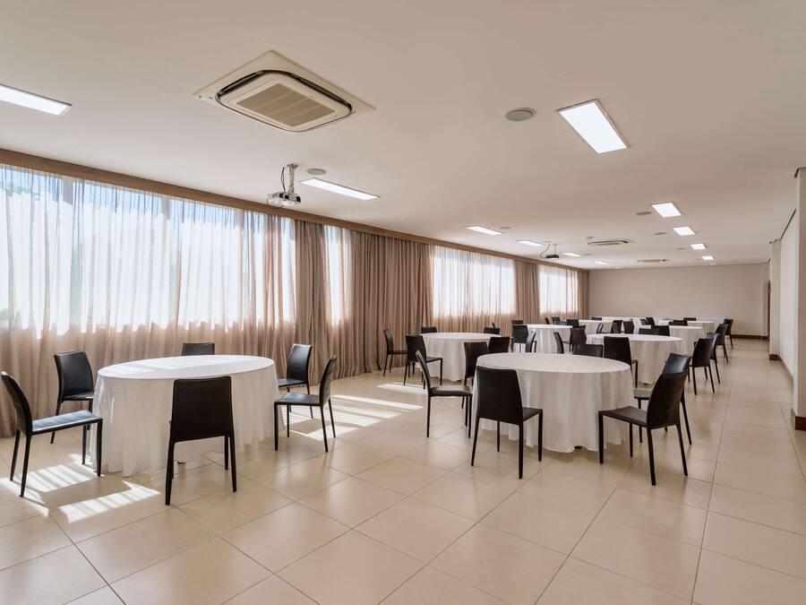 A conference room with multiple round tables and chairs, large windows with curtains, and ceiling lights.