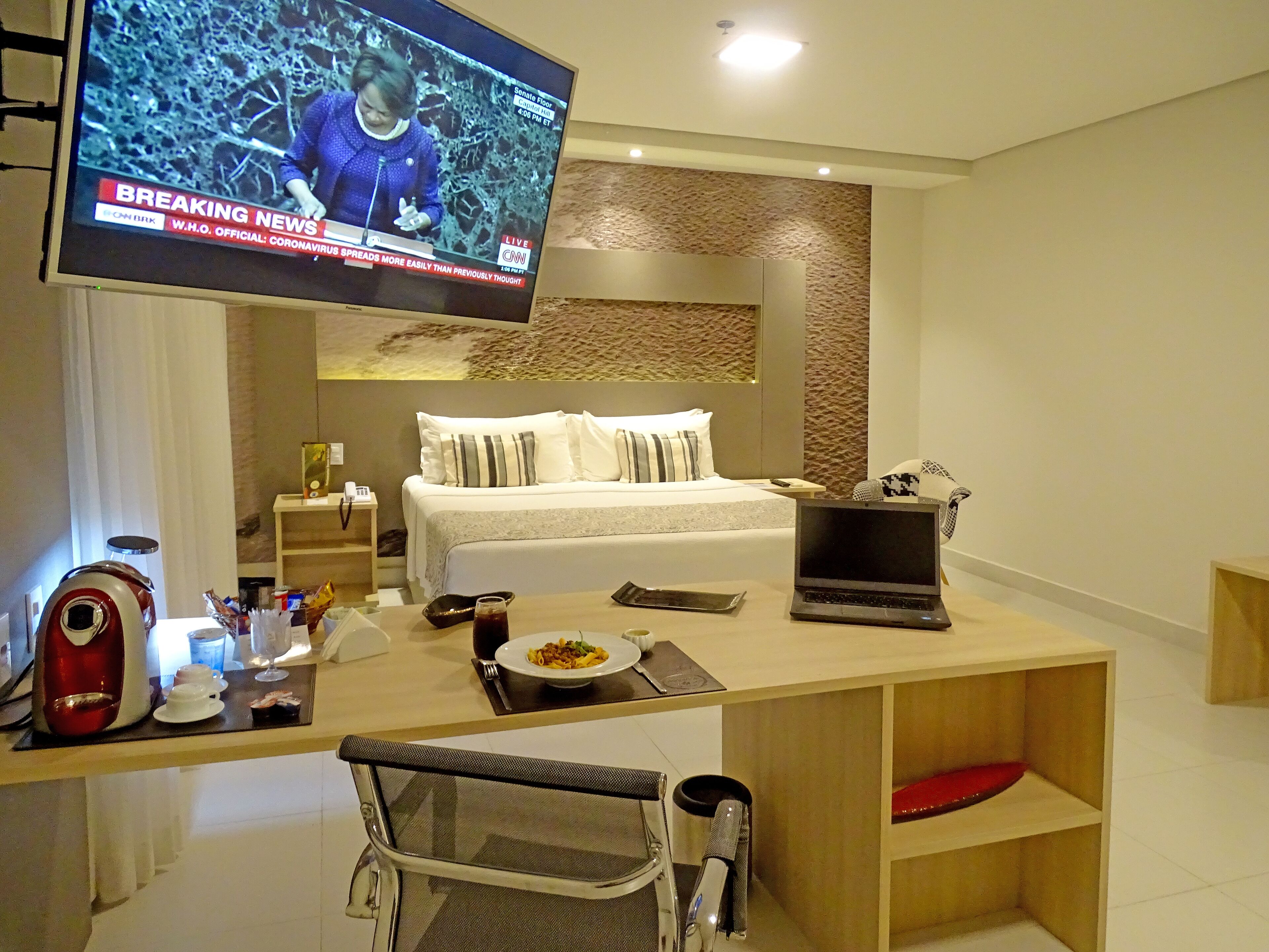 Quarto de hotel com cama, secretária com um laptop, televisão exibindo um noticiário e área de refeições com um prato de comida.