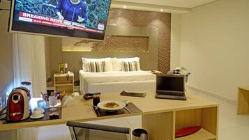Quarto de hotel com cama, secretária com um laptop, televisão exibindo um noticiário e área de refeições com um prato de comida.
