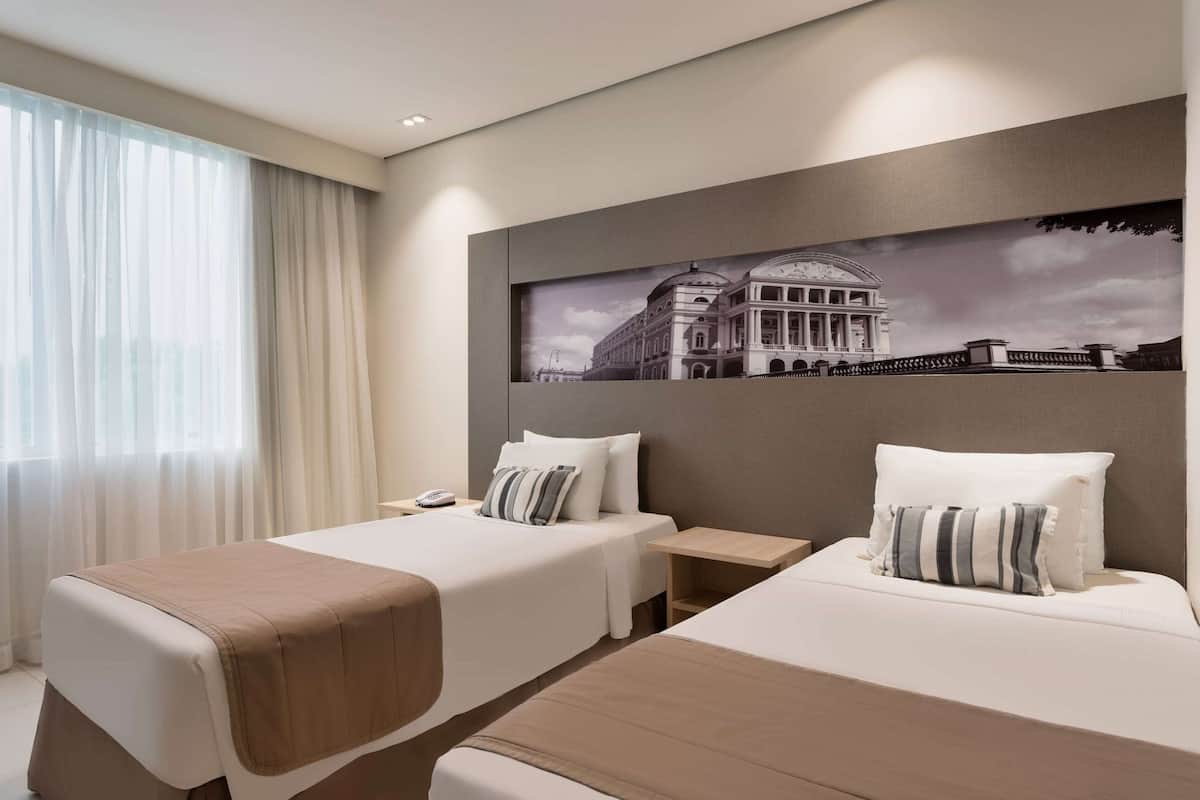 A hotel room with two beds, a large window with sheer curtains, a wall-mounted artwork of a building, and a bedside table with a lamp.