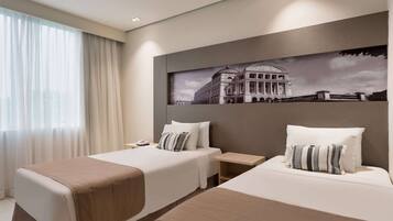 A hotel room with two beds, a large window with sheer curtains, a wall-mounted artwork of a building, and a bedside table with a lamp.