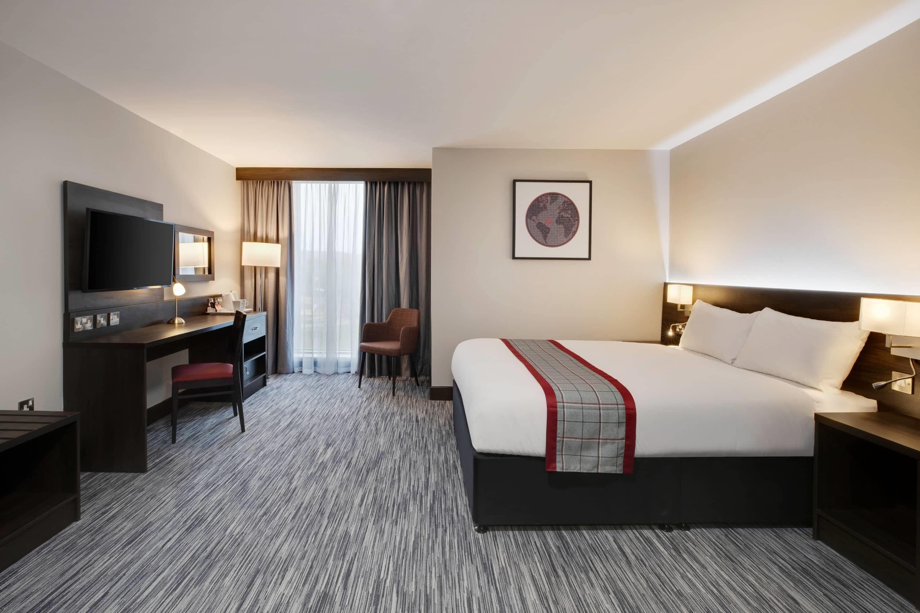 A hotel room with a bed, a desk, a chair, a TV, and a window with curtains.