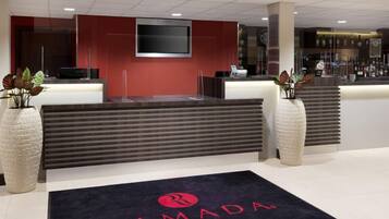 A reception area with a dark reception desk, a mounted TV, and a welcome mat with the logo "RAMADA".