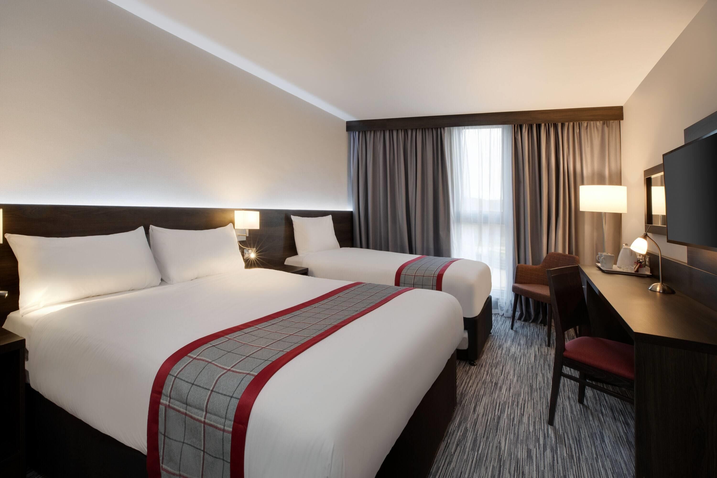A hotel room with two beds, a desk, a chair, a TV, and a window with curtains.