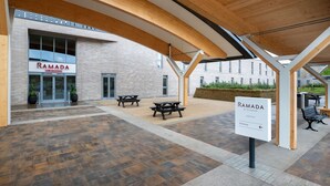 A covered outdoor area with picnic tables and benches, a sign for Ramada by Wyndham Leeds East, and a brick building in the background. - Ramada by Wyndham Leeds East (Leeds)