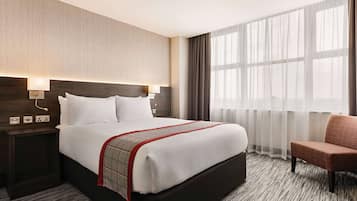 Una habitación de hotel con una cama grande, una mesita de noche con lámpara, una silla y una ventana con cortinas.
