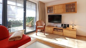 A living room with a sofa, a television, a cabinet with glass doors, and a view through a large window. - Papillon Whg. 12/4, Germany (Ostseebad Boltenhagen)
