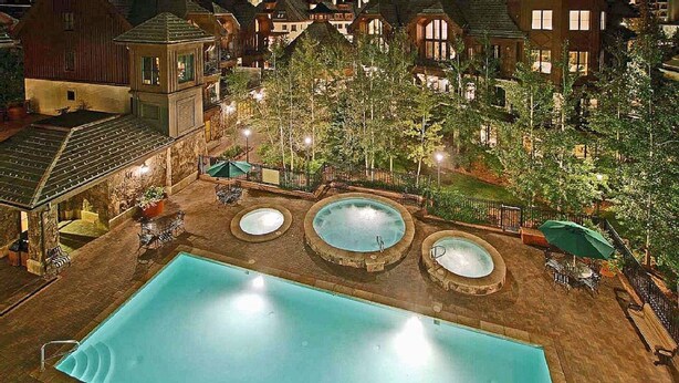 Una gran piscina al aire libre con tres jacuzzis, rodeada de árboles y un edificio de piedra.