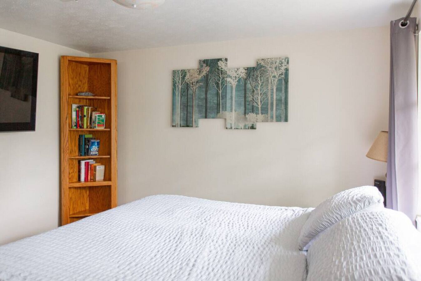 A bedroom with a bed, a wooden bookshelf, and two wall-mounted paintings.