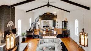 A spacious living area with a high ceiling, exposed beams, a fireplace, and a staircase leading to an upper level.