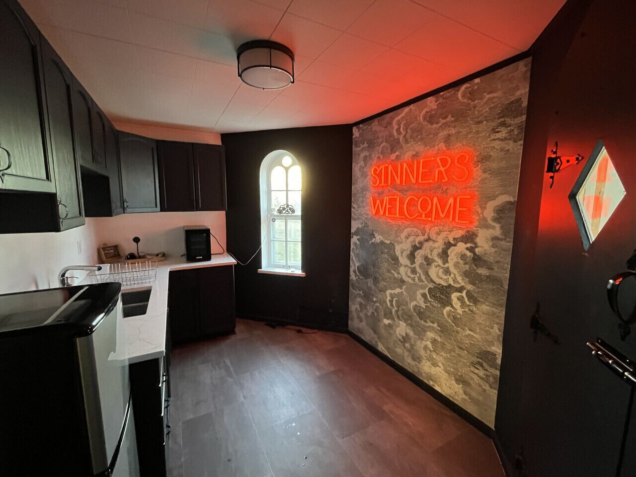 A modern kitchen with dark cabinets, a white countertop, and a neon sign reading "Sinners Welcome".