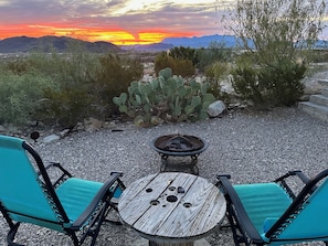 Un área de estar al aire libre con una mesa redonda y dos sillas, una chimenea y vistas a un paisaje desértico con montañas al fondo.