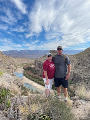 Dos personas en terreno rocoso, con un río y montañas al fondo.