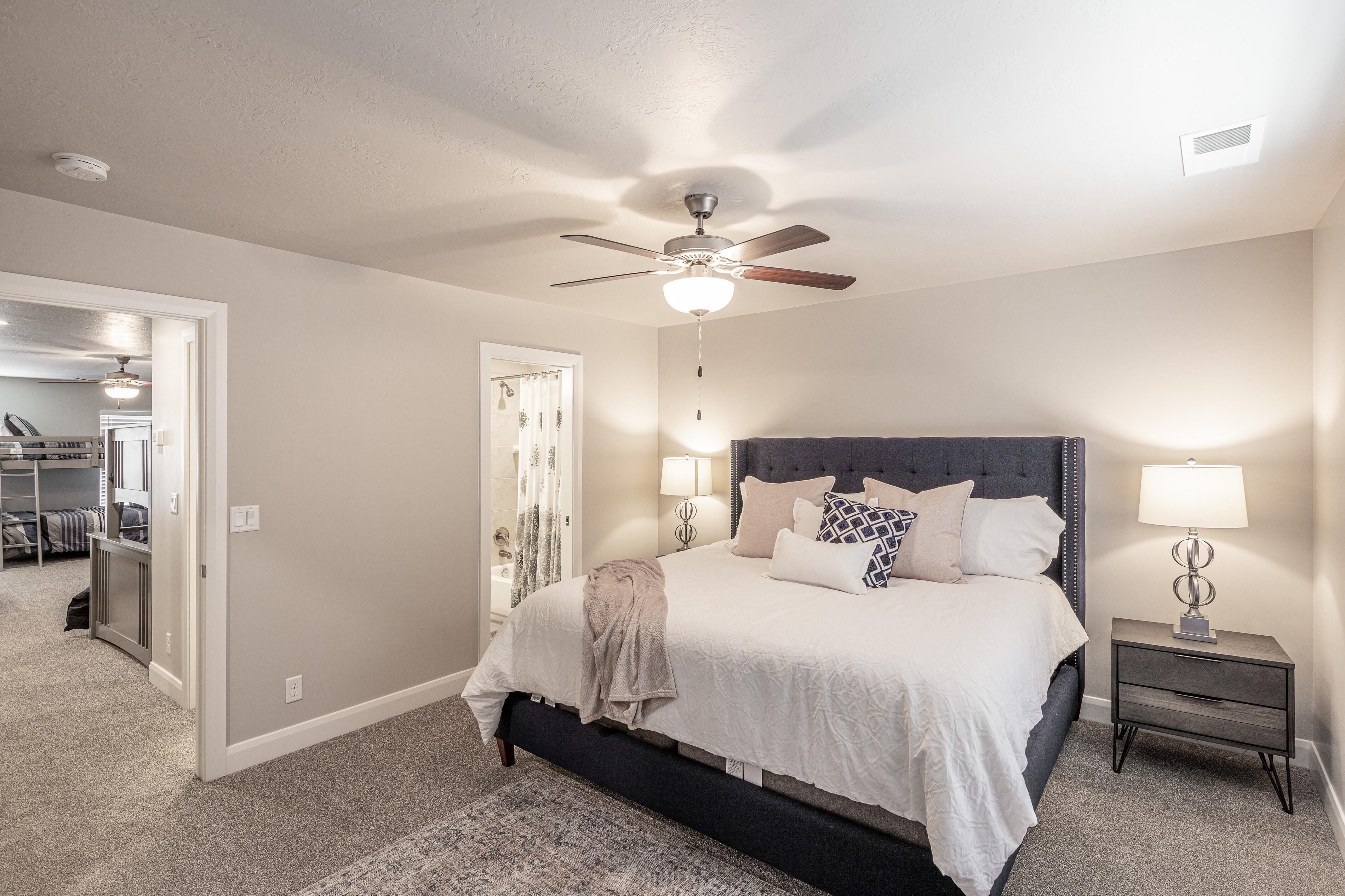 A bedroom with a bed, bedside lamps, a ceiling fan, and a door leading to another room.