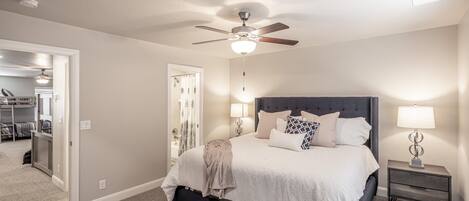 A bedroom with a bed, bedside lamps, a ceiling fan, and a door leading to another room.