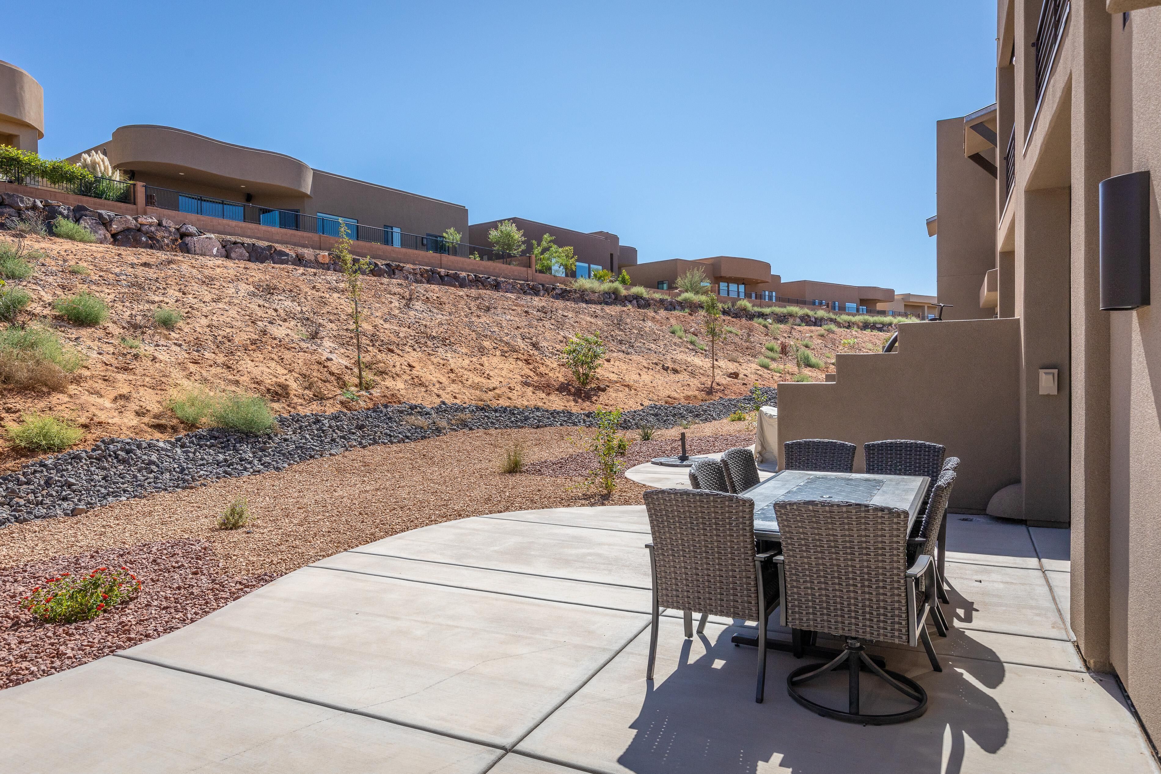 A patio area with a table and chairs, a landscaped garden with rocks and small plants, and a hillside with buildings in the background.