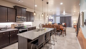 A modern kitchen with dark cabinets, stainless steel appliances, and a granite countertop. There is a dining area with a table and chairs, and a living room with a sofa and a TV. - Launchpad (St. George)