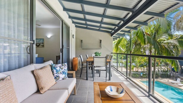 A modern living room with a sofa, dining table, and chairs, a large window, and a view of a swimming pool. - Baden 74 - Rainbow Shores. Top Floor - Air conditioned Unit - Walk To Beach (RAINBOW BEACH)