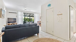 A living room with a sofa, TV, and a dining area with chairs. - Baden 74 - Rainbow Shores. Top Floor - Air conditioned Unit - Walk To Beach (RAINBOW BEACH)