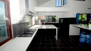 A modern kitchen with a central island, black tiled flooring, and a view of greenery outside. - Lausikula Chambres d'hôtes (Wallis Island)