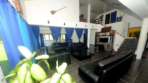 A spacious living area with a black leather sofa, a coffee table, and a TV. There is a staircase leading to an upper level, and blue curtains are visible. - Lausikula Chambres d'hôtes (Wallis Island)