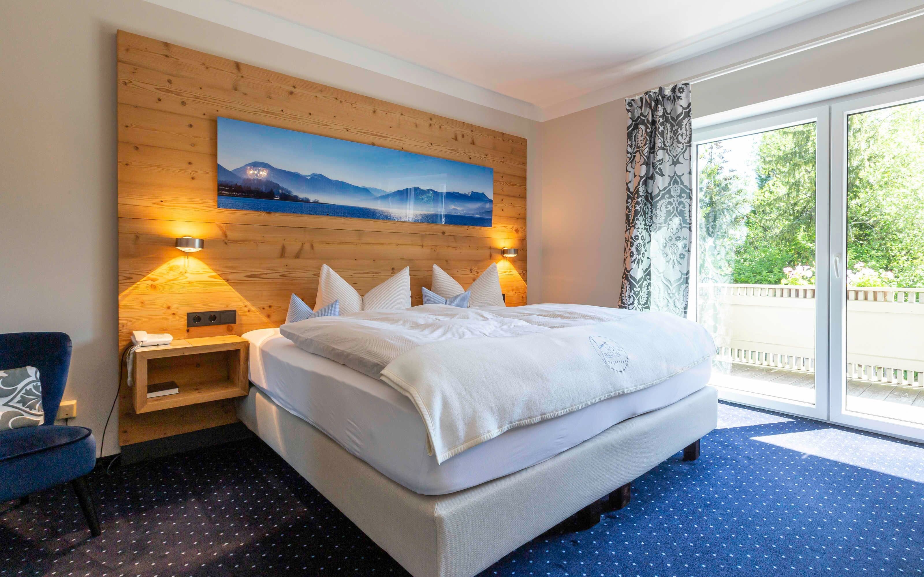 Habitación de hotel con cama, dos lámparas de noche, cabecera de madera, un cuadro, una silla y vistas a zonas verdes a través de una ventana.