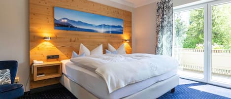 Habitación de hotel con cama, dos lámparas de noche, cabecera de madera, un cuadro, una silla y vistas a zonas verdes a través de una ventana.