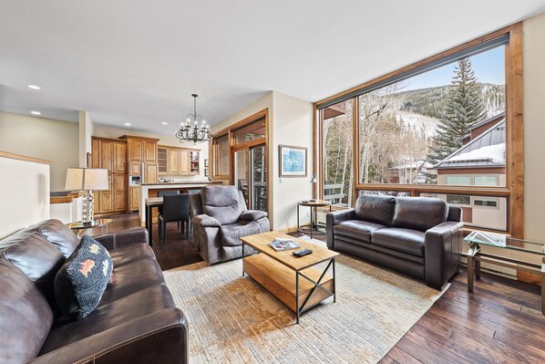 A spacious living room with a fireplace, leather sofas, a dining area, and a large window offering a view of a snowy mountain.