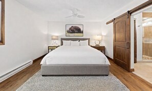 A bedroom with a large bed, two bedside tables with lamps, a ceiling fan, a wooden door, and a framed picture on the wall.