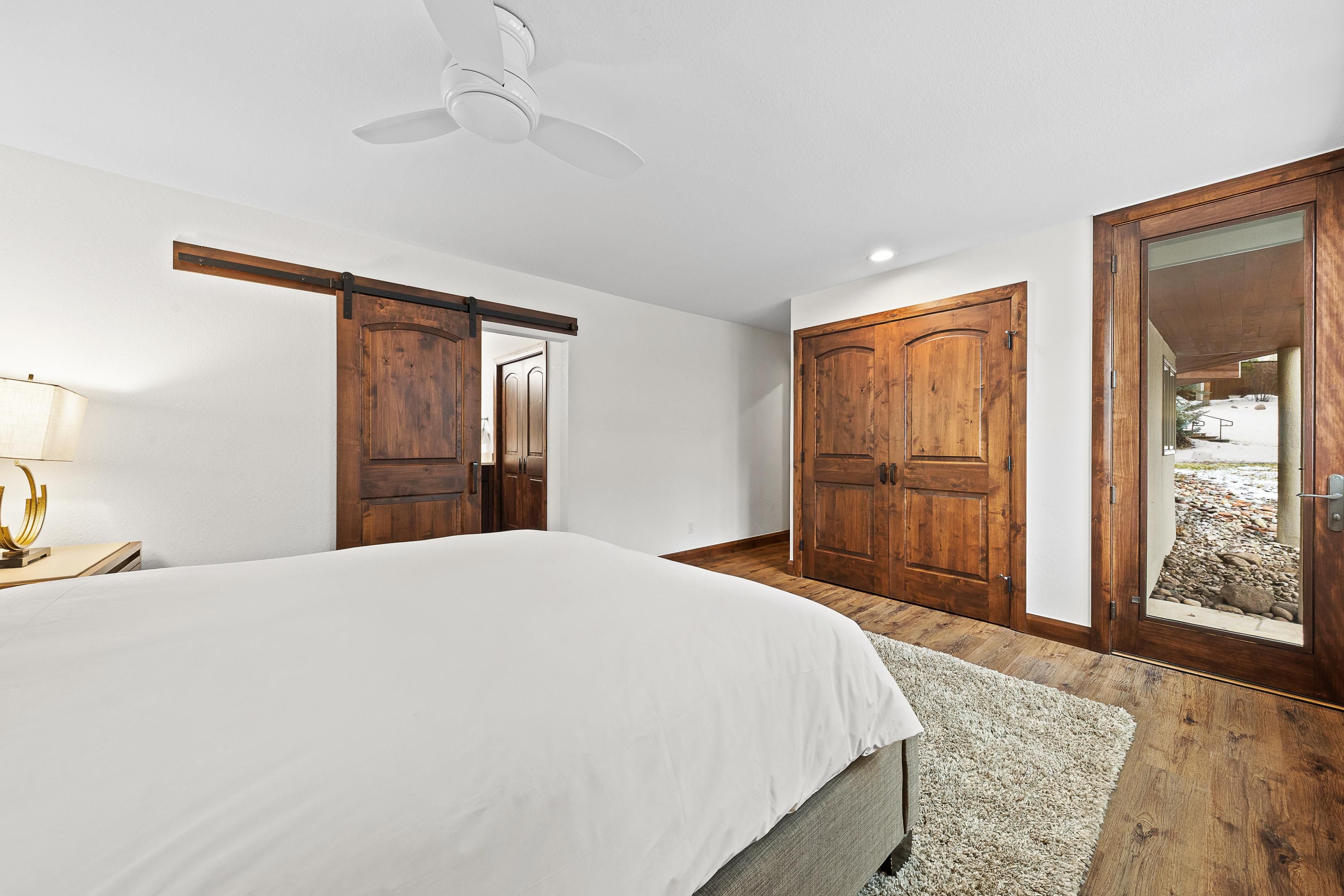 A bedroom with a bed, a ceiling fan, wooden doors, and a view through a glass door.