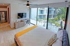 A modern bedroom with a large bed, a desk, and a TV. There is a balcony with a view of buildings and greenery.